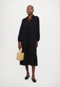 Vestido longo preto com um decote com atacadores, mangas longas e barra em camadas. Combinado com uma mala de palha amarela e sapatos pretos rasos.