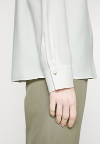 Blouse vert clair avec une texture lisse, manches longues et détails de boutons au niveau des poignets, associée à un pantalon beige ajusté.