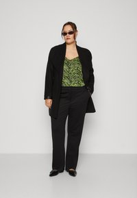 Black oversized coat over a green and black zebra-striped top, paired with black pants and flat pointed shoes. Stylish sunglasses complete the look.