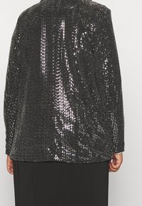 Giacca nera con paillettes che riflette la luce, indossata da una persona vista di spalle con le mani visibili ai lati su uno sfondo semplice.