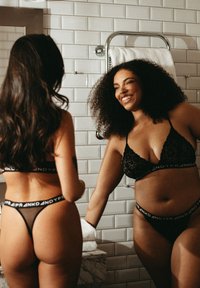 Zwart lingeriesetje met kanten bralette en doorzichtige string, afgewerkt met merk-elastische banden. De achtergrond bestaat uit witte tegels en opgevouwde handdoeken.