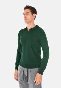 Polo sweater verde a maniche lunghe con scollo a V e polsini a coste, abbinato a pantaloni grigio chiaro con cordini e vestibilità ampia.