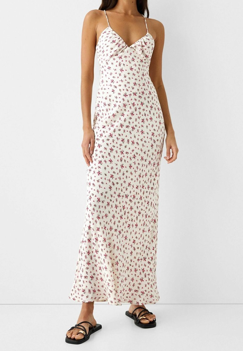 Femme portant une robe-jupon blanche longue jusqu'aux chevilles, avec des fines bretelles et un petit motif floral rouge, associée à des sandales plates noires.