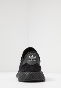 Svart Adidas-sneaker med övre nät, texturerad gummisula, rundad form och logga på hälen. Minimalistisk design och strömlinjeformad silhuett.