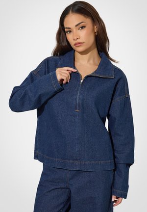 Mujer con pelo largo y castaño lleva una chaqueta vaquera azul oscuro con cremallera y pantalones vaqueros a juego sobre un fondo neutro.