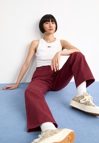 Top alb cu textul "OBEY", pantaloni visinii cu talie înaltă și încălțăminte texturată bej. Modelul stă pe un fundal albastru.