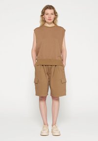 Bruine mouwloze top gecombineerd met bruin cargoshorts tot de knie; beide hebben een ontspannen pasvorm en eenvoudige stikdetails. Beige sneakers maken de look af.