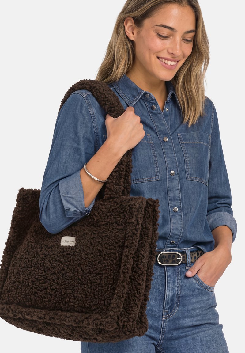 Femme en chemise en denim et jean tenant un grand sac cabas marron foncé texturé sur l'épaule, souriante et regardant vers le bas.