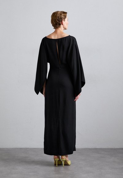 Black long-sleeve dress with a fitted silhouette, open back detail, and flowing sleeves. Paired with yellow high-heeled shoes.