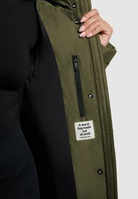 Olivovo zelená prešívaná bunda s čiernou vnútornou podšívkou, bočnou zipsovou kapsou a štítkom s nápisom "Teplé objatie vyrobené z látky."