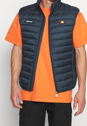 Mann trägt eine marineblaue gesteppte ärmellose Weste über einem hellorangefarbenen kurzärmeligen Hemd und schwarzen Cargohosen, steht mit entspannten Armen.