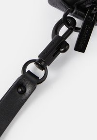 Črn usnjen trak z mat površino, opremljen z kovinsko zaponko in dodatki z našitim logotipom. Vključuje okroglo zanko za pritrjevanje.