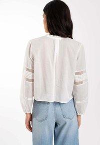 Blusa blanca, ligera, con mangas largas, detalles de encaje texturizado en las mangas y un plisado en la parte posterior. Combinada con vaqueros azules de tiro alto.