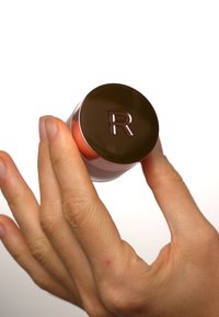 Rosé zylindrische Flasche, die in einer Hand gehalten wird, mit einem glänzenden, dunkelbraunen Deckel, der mit dem eingeprägten Buchstaben "R" verziert ist. Glatte Oberfläche, minimalistisches Design.