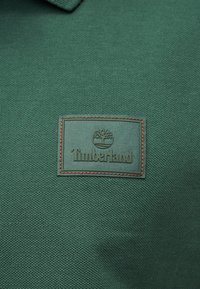 Πράσινο υφασμάτινο ύφασμα με υφασμένη ορθογώνια κηλίδα που φέρει το λογότυπο Timberland και το έμβλημα δέντρου στο κέντρο.