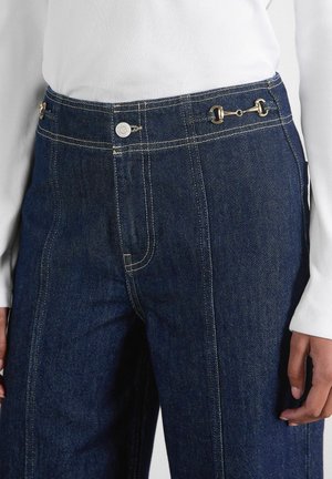 Denim jeans with a dark blue color, high waist, gold hardware accents, and contrasting stitching details. Features side pockets and a button closure.
