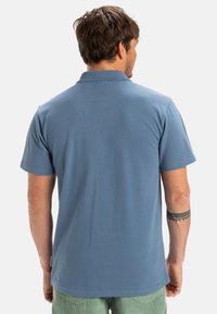 Blå bomullspolo med klassisk krage och korta ärmar, med rak nederkant och liten etikett på sidan.