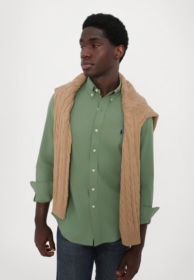 Chemise boutonnière vert clair à manches longues, associée à un pull en maille beige drapé sur les épaules. Jean en denim foncé.