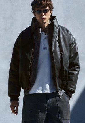 Jeune homme portant une veste en cuir noire, une chemise blanche, un pantalon sombre et des lunettes de soleil, debout devant un fond gris clair uni.