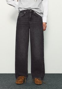 Osoba nosiaca voľné čierne rifle s vysokým pásom, hnedé mokasíny, sivé tričko a bielu košeľu s dlhým rukávom, stojaca na šedom koberci.