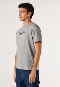 Tricou din bumbac gri cu mâneci scurte, având un logo roșu brodat pe față. Design standard cu guler rotund.