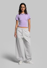 Lilafarbnes, eng anliegendes T-Shirt mit kurzen Ärmeln, kombiniert mit übergroßen weißen Sweatpants mit einem dunklen Logo und weißen Turnschuhen. Einfacher, lässiger Look.