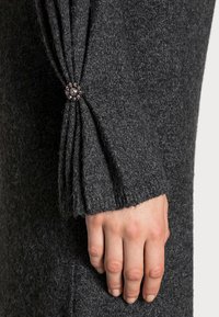 Pull gris foncé avec un tissu texturé. Le poignet présente des plis maintenus par un bouton décoratif avec des perles. Une main est partiellement visible, reposant.