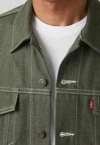 Olivgrön denimjacka med vit sömnad, metallknappar, framfickor och en röd Levi's-logotyp på den nedre sidan.