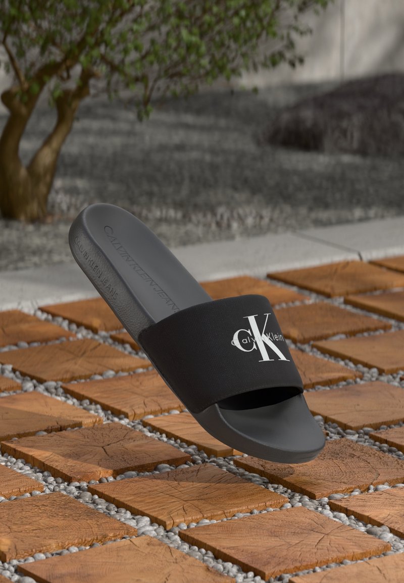 Sandale slide noire Calvin Klein avec logo blanc sur la bride, affichée au-dessus d'une dalle en bois et d'un chemin de gravier à l'extérieur près d'un arbre.