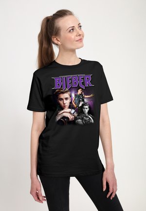 Kvinde med langt hår iført en sort t-shirt med tre billeder af Justin Bieber og navnet "Bieber" i lilla bogstaver.