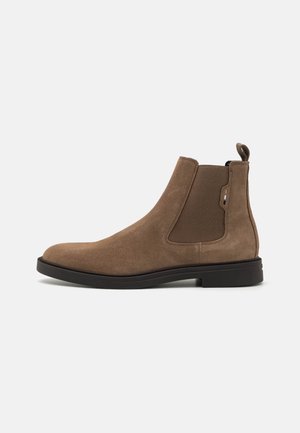 Bruine suède Chelsea-boot met een elastische zijpaneel en zwarte zool op een witte achtergrond, zijaanzicht.