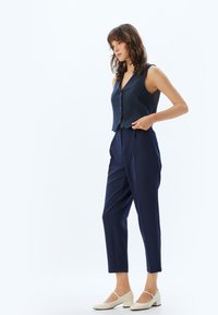 Marineblauwe geribbelde vest met knopen en op maat gemaakte marineblauwe broek. Lichtgekleurde blokhak schoenen. Minimalistisch ontwerp met een gladde textuur.