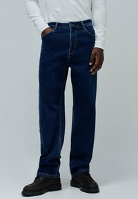 Jeans de mezclilla azul oscuro con un corte relajado, que cuentan con cinco bolsillos y costuras visibles. Llevados con zapatos negros de suela plana.