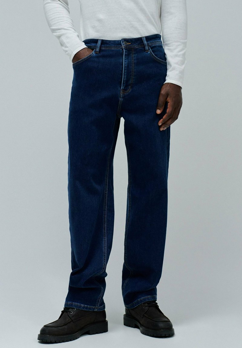 Jeans de mezclilla azul oscuro con un corte relajado, que cuentan con cinco bolsillos y costuras visibles. Llevados con zapatos negros de suela plana.