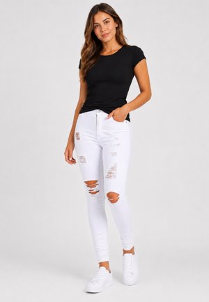 Kvinde står iført en sort kortærmet top, revnede hvide skinny jeans og hvide sneakers på en ensartet lys baggrund.