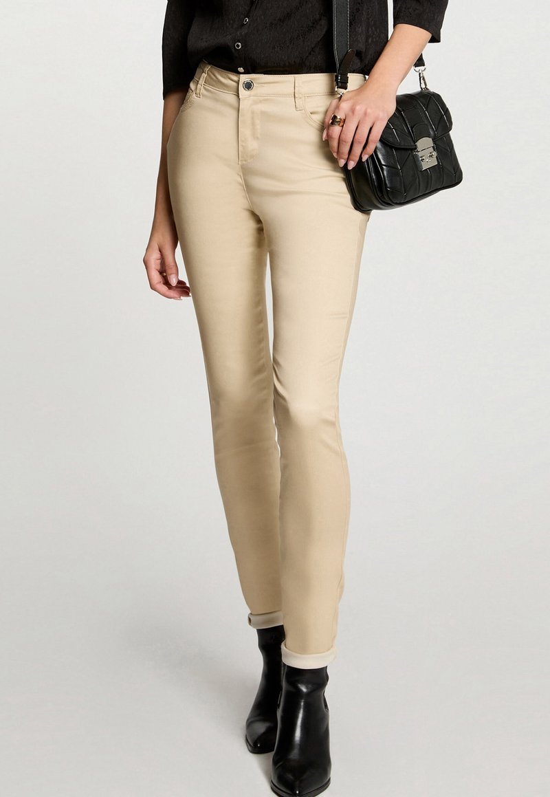 Femme portant un pantalon slim beige, un haut noir boutonné, des bottines noires, portant un sac à main matelassé noir avec une boucle argentée.