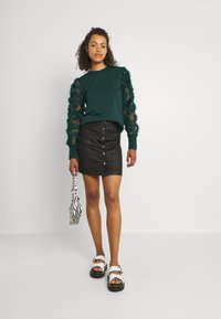 Dark green sweater with floral mesh sleeves, paired with a black button-front mini skirt and white sandals. Accented with a zebra print bag.