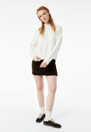 Witte, gebreide trui met hoge hals, gecombineerd met een bruine, gestructureerde minirok. Het model draagt witte kniehoge sokken en bruine sneakers.