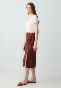 T-shirt bianco a scollo a V abbinato a una gonna midi marrone con uno spacco laterale e un dettaglio annodato in vita. Sandali flat marroni.