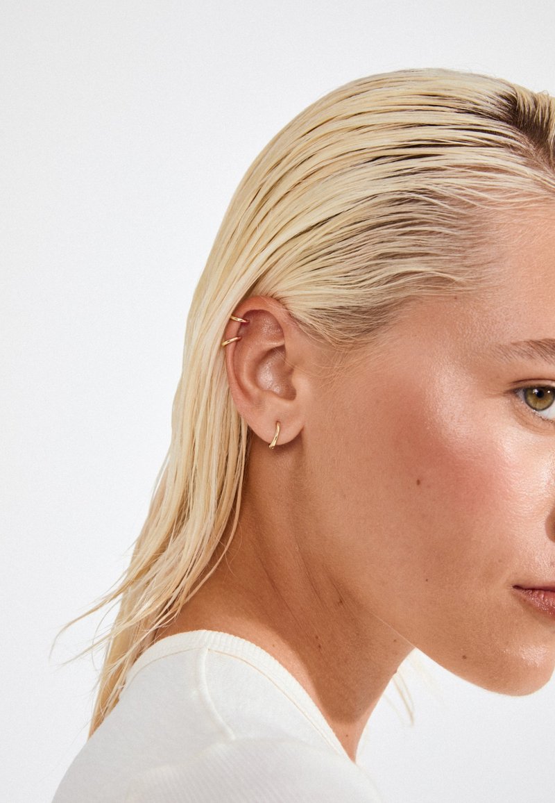 Zlaté kruhové náušnice vystavené na uchu modelky, s elegantním designem a lesklým povrchem. Vlasy jsou zpěněné dozadu, což odhaluje několik píchání do uší.