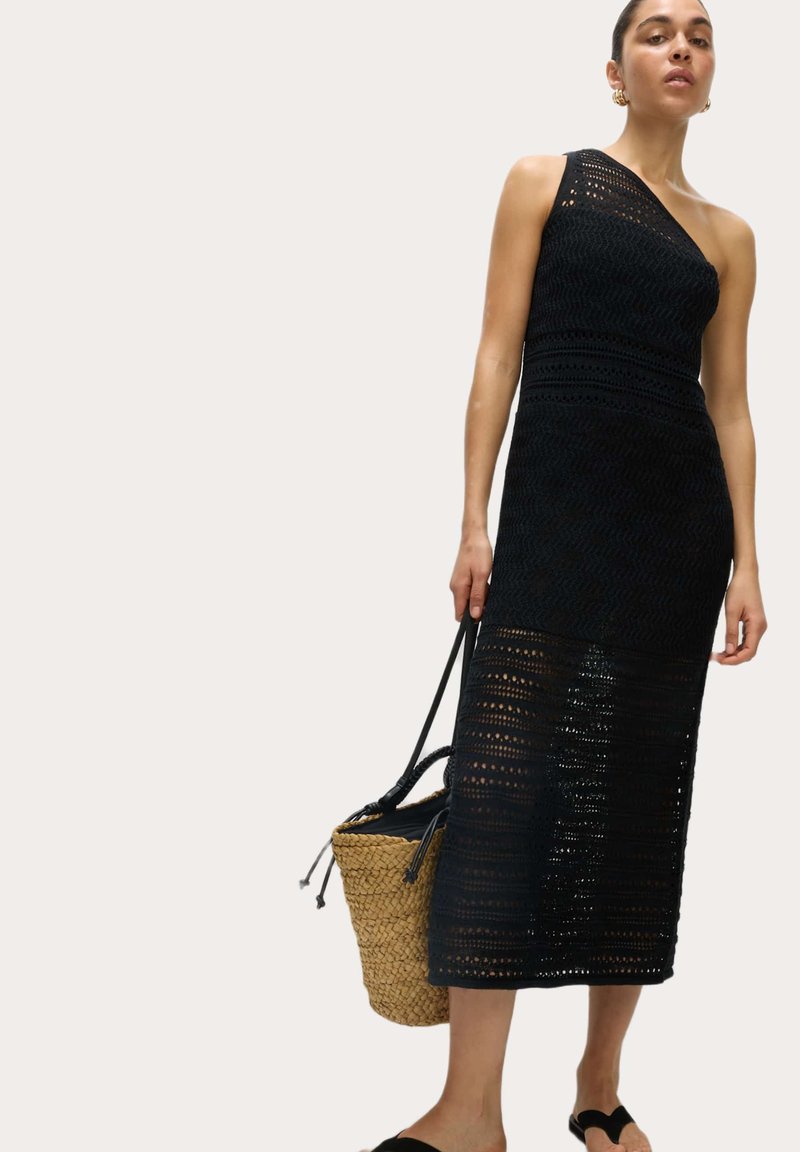 Robe en tricot noire à une épaule, mettant en avant un design texturé en tricot ouvert avec une silhouette droite et une longueur mi-mollet. Le modèle tient un sac tissé.