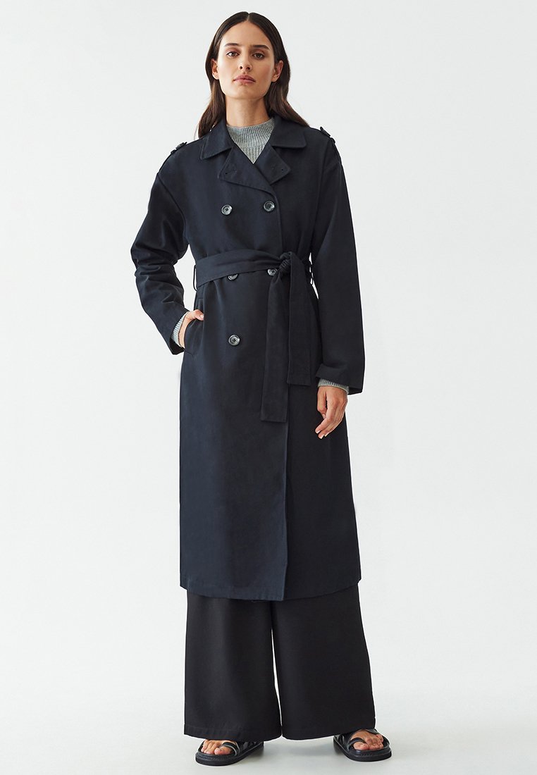 Tumesinine pikk trench coat, kahekordsete nööpidega, vöökoha pealt talje ja sisselõigatud kraega. Valmistatud siledast kangast, millel on struktureeritud sobivus.