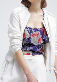 Kvinna iklädd en vit zip-up jacka över en blå blommig och röd fiskmönstrad volangblus samt vita höga byxor.