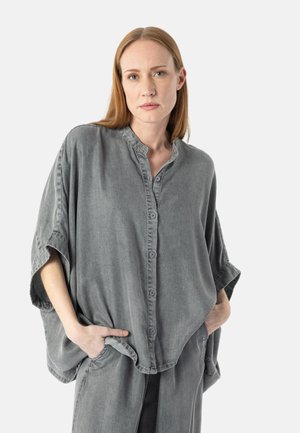 Woman with straight red hair wearing loose, washed gray button-up shirt and matching wide-leg pants, hands in pockets, neutral expression.