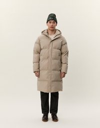 Uomo che indossa un lungo cappotto imbottito beige con cappuccio, pantaloni neri, scarpe marroni scure e un berretto verde scuro su uno sfondo chiaro e semplice.