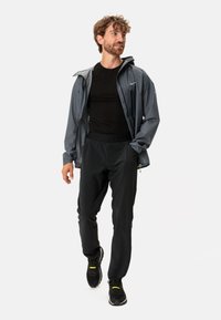 Giacca grigia impermeabile da uomo sopra una maglietta nera aderente, abbinata a pantaloni neri a sigaretta e sneaker neri con accenti gialli.
