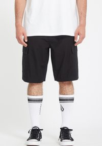 Schwarze Cargo-Shorts mit seitlichen Taschen, getragen mit weißen Kniestrümpfen mit schwarzen Streifen und schwarzen Sneakers mit weißen Sohlen.