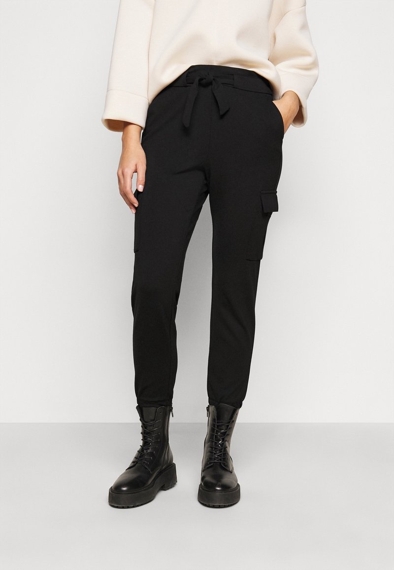 Personne portant un pantalon cargo noir avec une ceinture à nouer, associé à des bottes de combat noires lacées et un pull oversize crème.