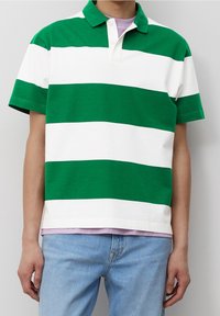 Henkilö, jolla on päällään vihreä ja valkoinen leveäraitainen lyhythihainen polo-paita vaaleanliilan paidan päällä, ja vaaleansiniset farkut yksinkertaista taustaa vasten.