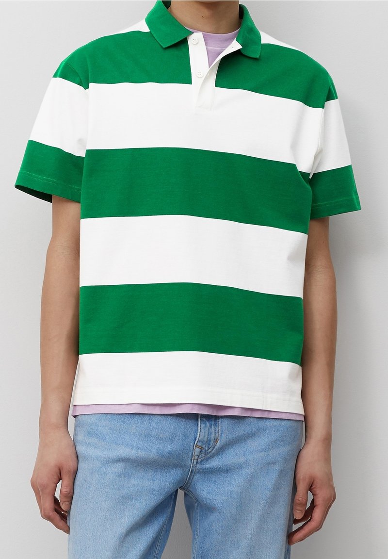 Henkilö, jolla on päällään vihreä ja valkoinen leveäraitainen lyhythihainen polo-paita vaaleanliilan paidan päällä, ja vaaleansiniset farkut yksinkertaista taustaa vasten.